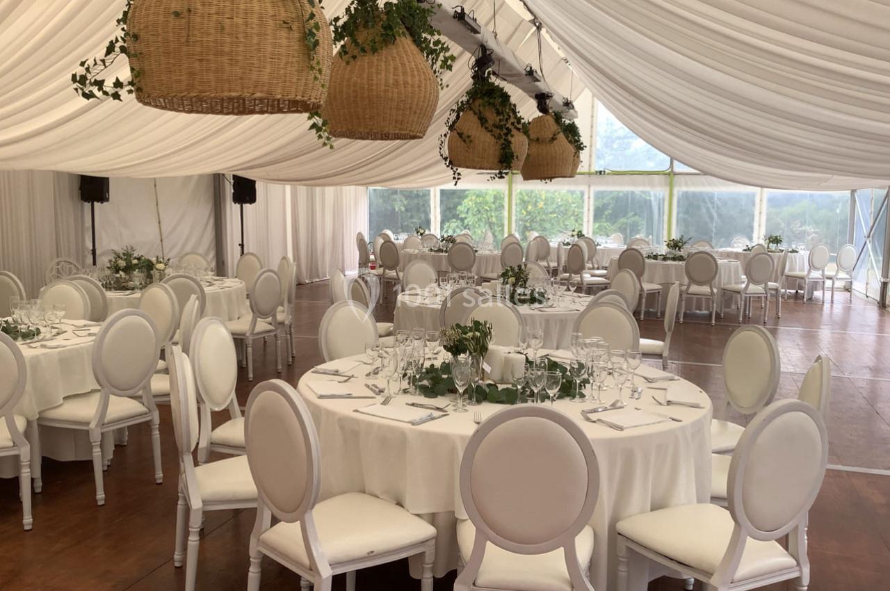 Salle de réception sous chapiteau avec tables rondes dressées, chaises blanches et suspensions en osier décorées de verdure.