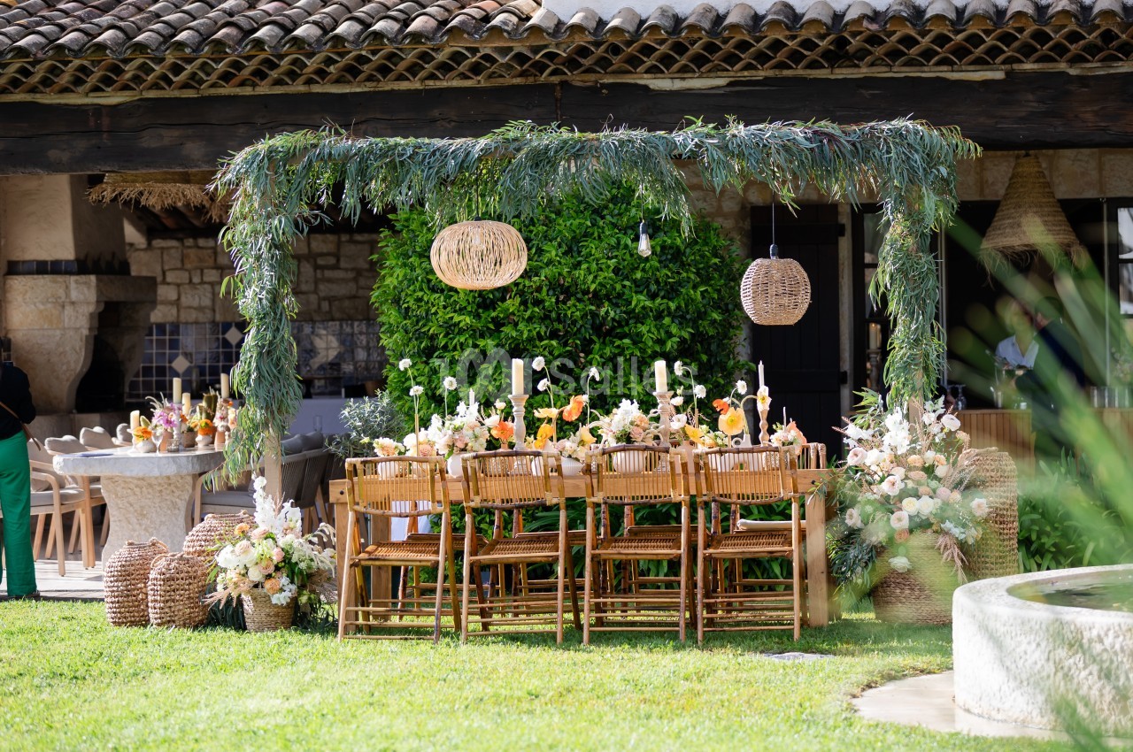 Table en bois décorée de fleurs et bougies sous une pergola ornée de feuillage, dans un jardin ensoleillé.