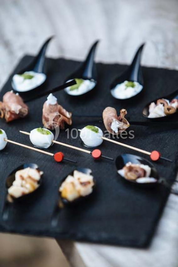 Assortiment de bouchées apéritives sur un plateau noir, comprenant brochettes et cuillères garnies.