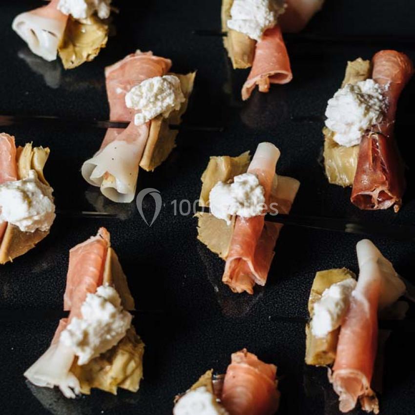 Assortiment de bouchées sur des pics, composées de charcuterie, fromage frais et artichauts, disposées sur une surface noire.