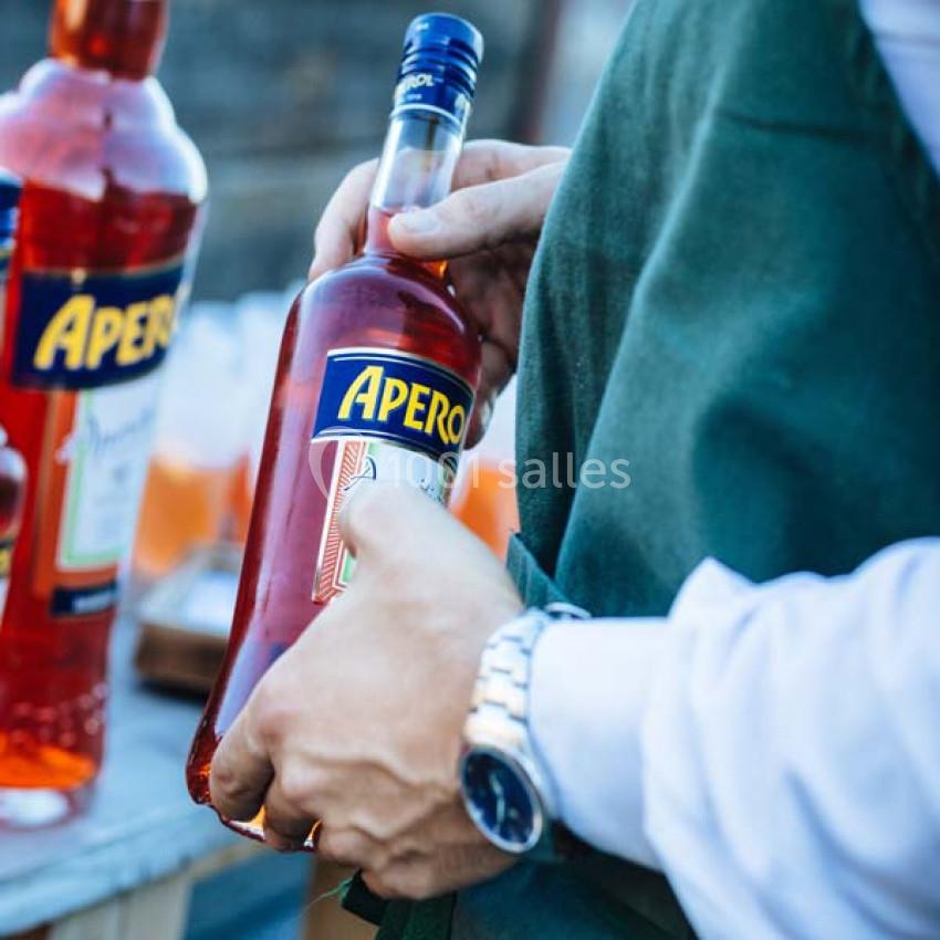 Une personne tenant une bouteille d'apéritif rouge avec une étiquette ’Apero’ lors d'un événement en extérieur.