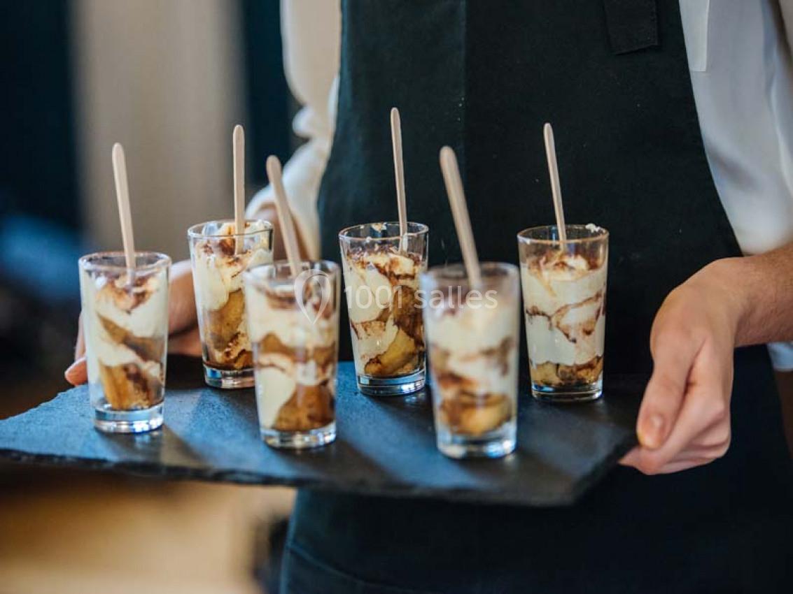Six verrines de tiramisu sont présentées sur un plateau noir, tenues par une personne en tablier.