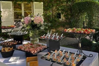 Buffet en extérieur avec divers plats et amuse-bouches disposés sur des présentoirs, entouré de verdure et de fleurs.