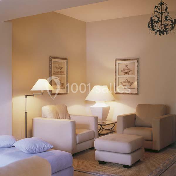 Deux fauteuils beiges, un pouf et des lampes dans un salon éclairé, décoré de cadres et d'un lustre noir.