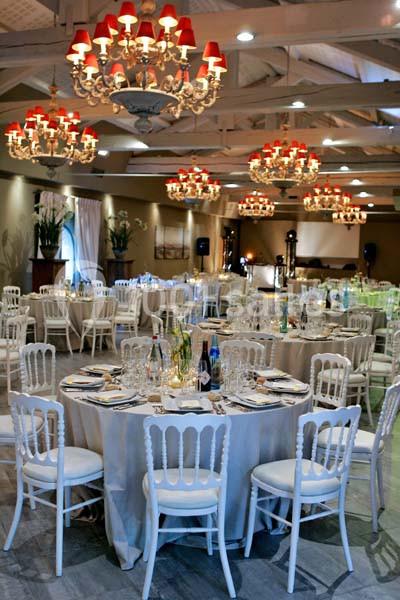 Salle de réception élégante avec tables rondes dressées, chaises blanches et lustres ornés de lampes rouges.