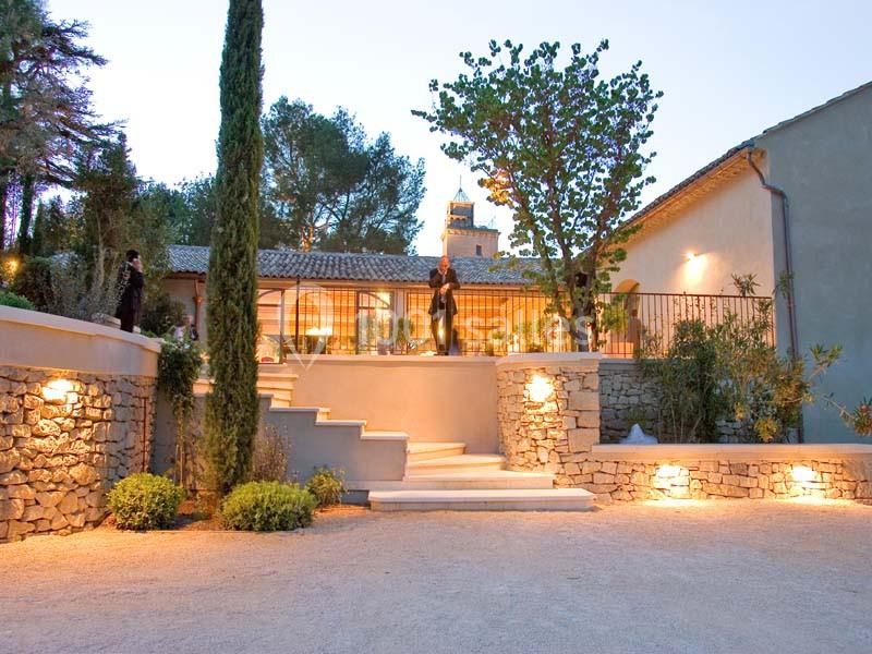 Façade d'une maison en pierre avec terrasse éclairée, entourée de végétation et d'arbres au crépuscule.