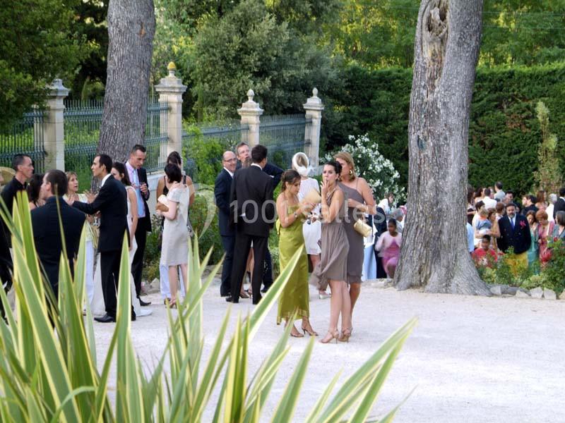 Groupe de personnes élégamment vêtues discutant dans un jardin avec des arbres et une allée gravillonnée.