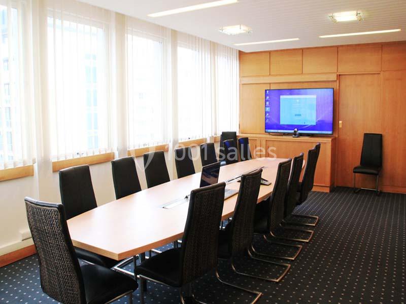 Salle de réunion lumineuse avec une grande table rectangulaire, des chaises noires et un écran mural allumé.