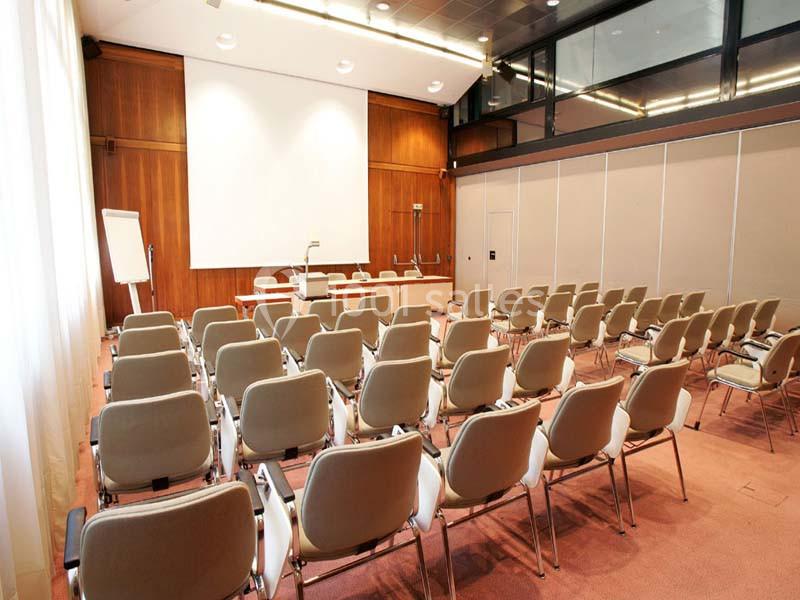 Salle de conférence vide avec des rangées de chaises beige, un écran de projection et un tableau blanc.