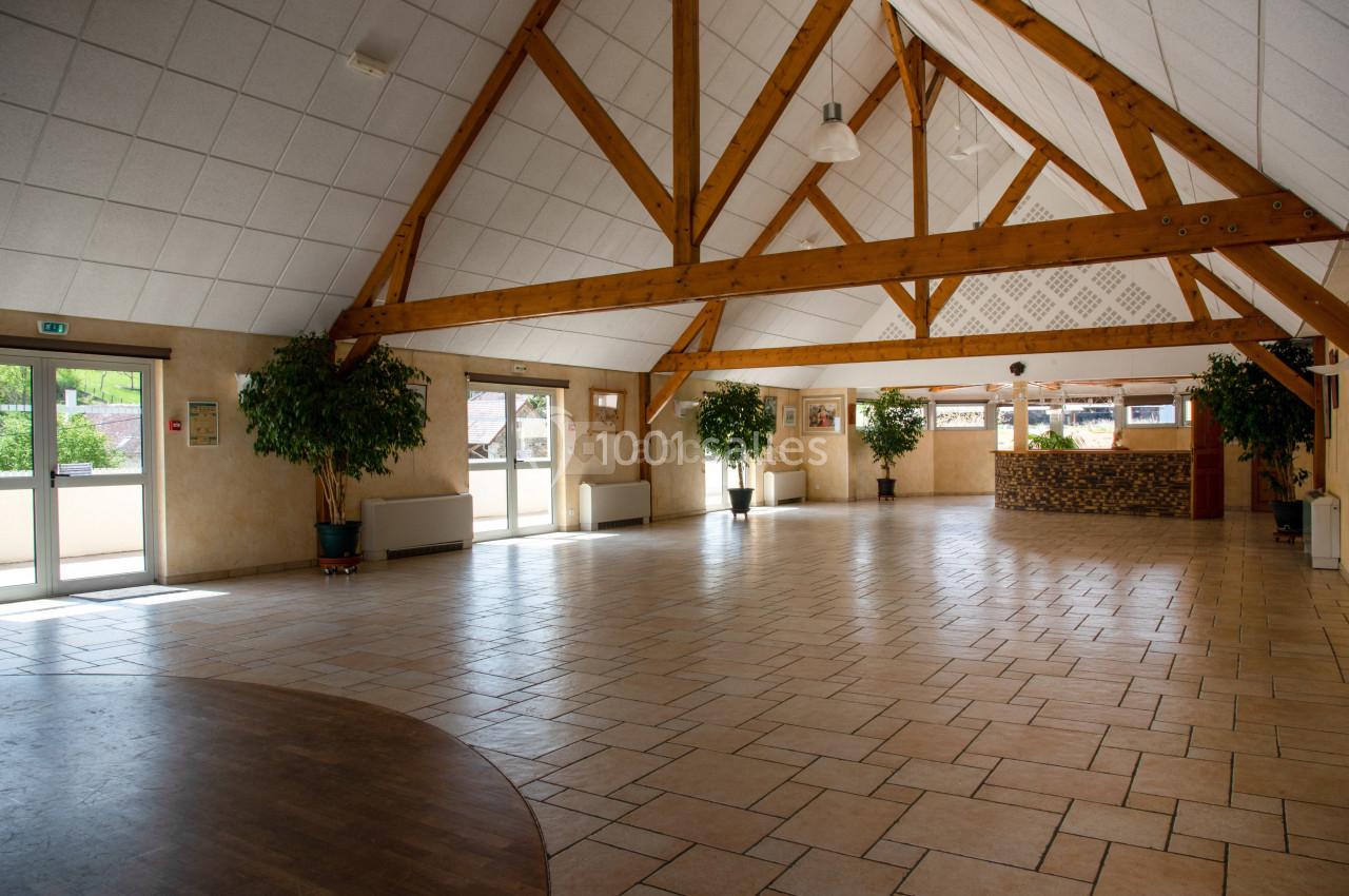 Salle spacieuse avec charpente en bois apparente, sol carrelé, grandes fenêtres et comptoir en pierre au fond.