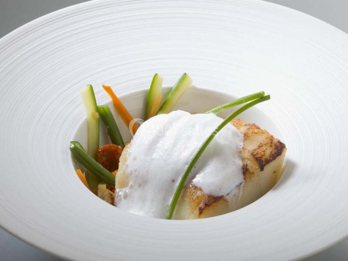 Plat gastronomique avec poisson grillé, légumes croquants et émulsion blanche, présenté dans une assiette creuse blanche.