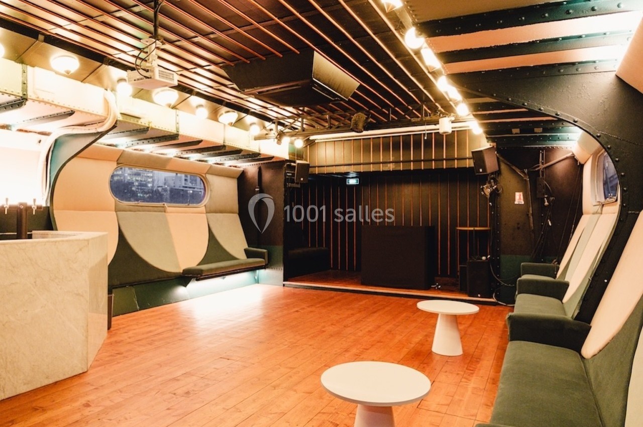 Salle intérieure moderne avec parquet, banquettes rembourrées, tables basses blanches et éclairage chaleureux.