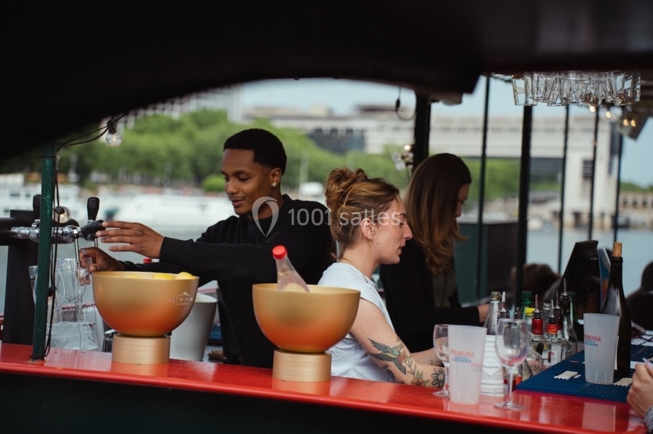 Des personnes préparent des boissons derrière un bar en extérieur, avec une rivière et des arbres en arrière-plan.