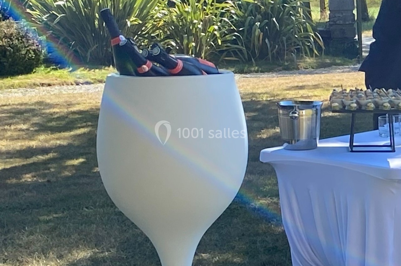 Bouteilles de champagne dans un grand seau en forme de verre, posées sur une pelouse près d'une table de réception.