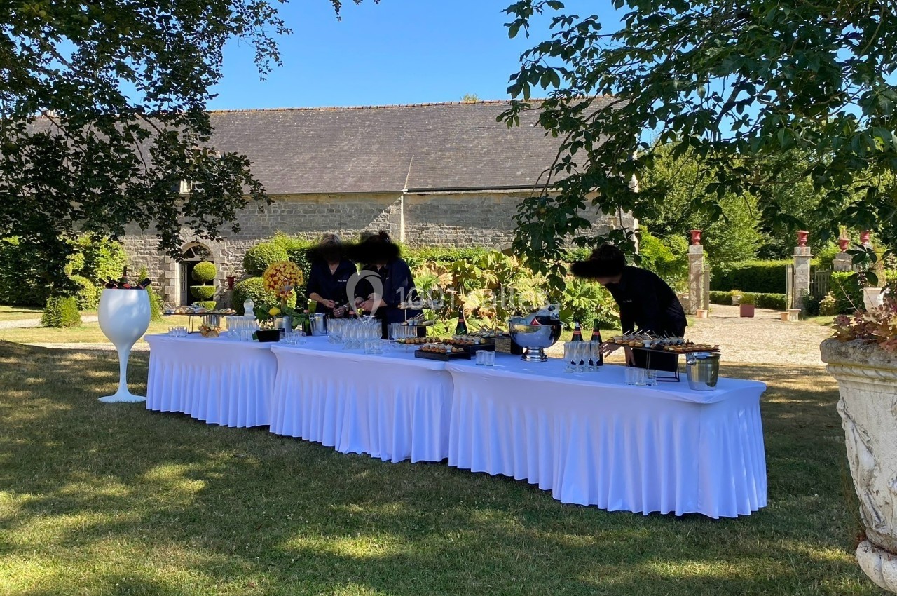 Table de buffet dressée en extérieur avec nappes blanches, entourée de verdure et d'un bâtiment en pierre en arrière-plan.