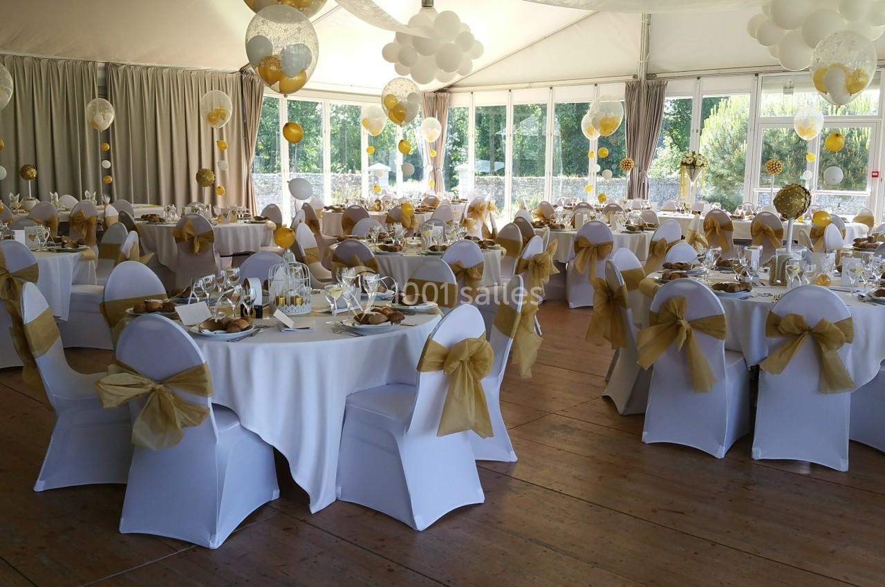 Salle de réception décorée avec des tables rondes, nappes blanches, chaises ornées de nœuds dorés et ballons suspendus.