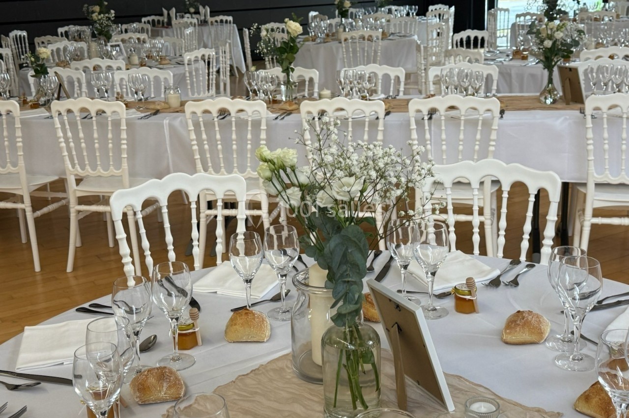 Salle de réception décorée pour un événement, avec tables dressées, nappes blanches, fleurs et couverts élégants.