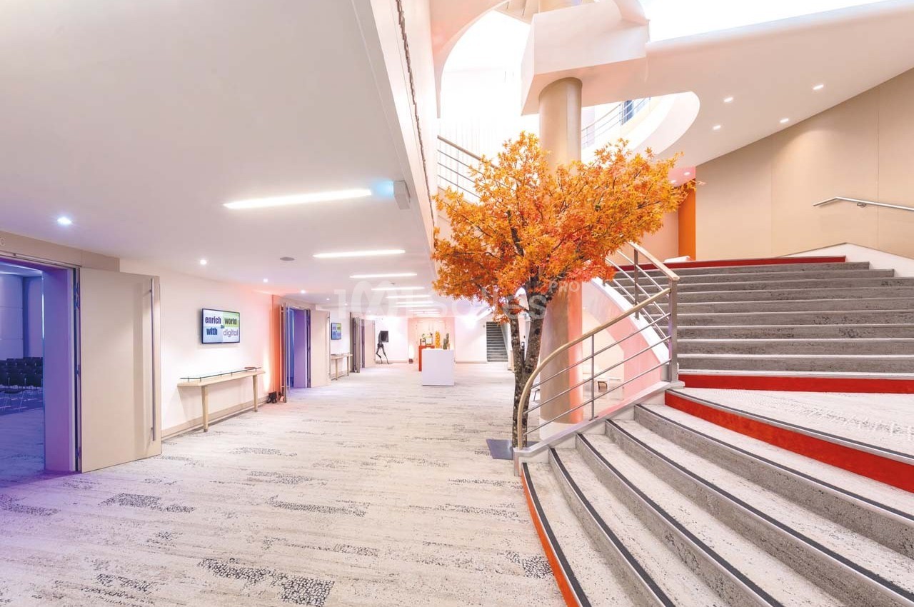 Hall lumineux avec escalier moderne, arbre décoratif orange et éclairage naturel provenant d'une verrière.