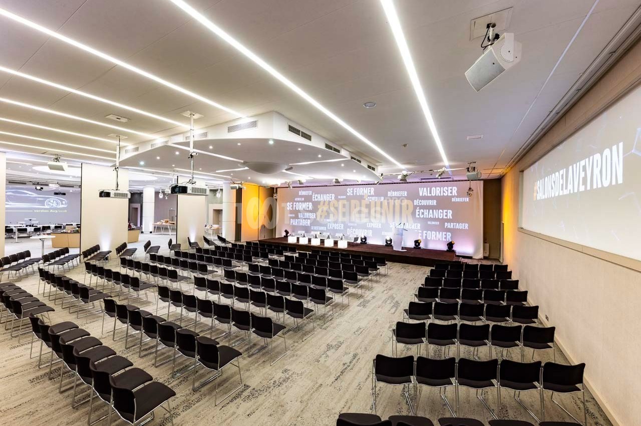 Salle de conférence moderne avec rangées de chaises noires, éclairage au plafond et écrans projetant des textes.