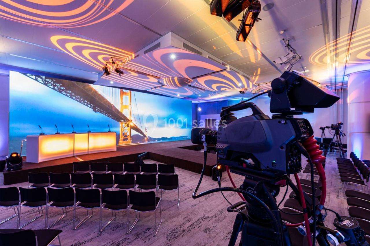 Salle de conférence équipée de caméras, éclairage coloré et scène avec pupitres devant un écran géant.