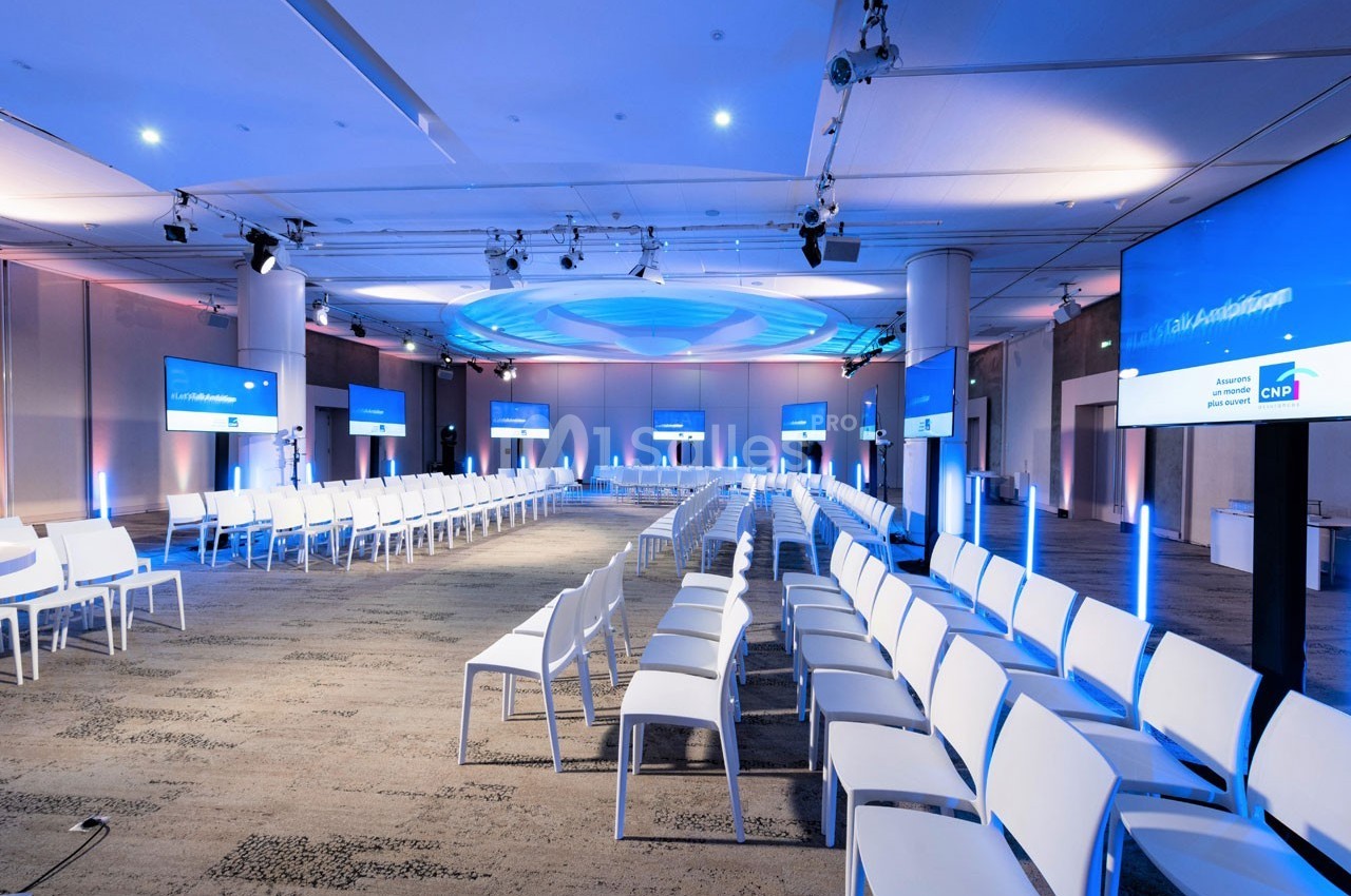 Salle de conférence moderne avec rangées de chaises blanches, écrans et éclairage bleu.