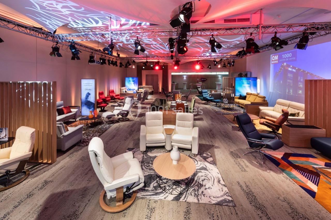 Salle d'exposition avec fauteuils et canapés disposés sur des tapis, éclairage coloré et projecteurs au plafond.