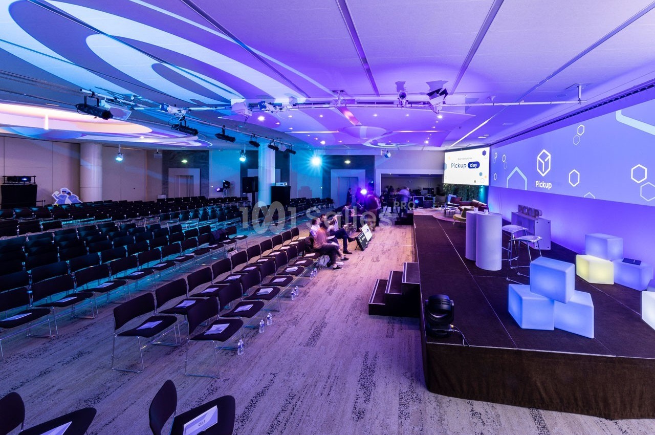 Salle de conférence moderne avec des sièges alignés, une scène équipée et des écrans affichant des visuels lumineux.