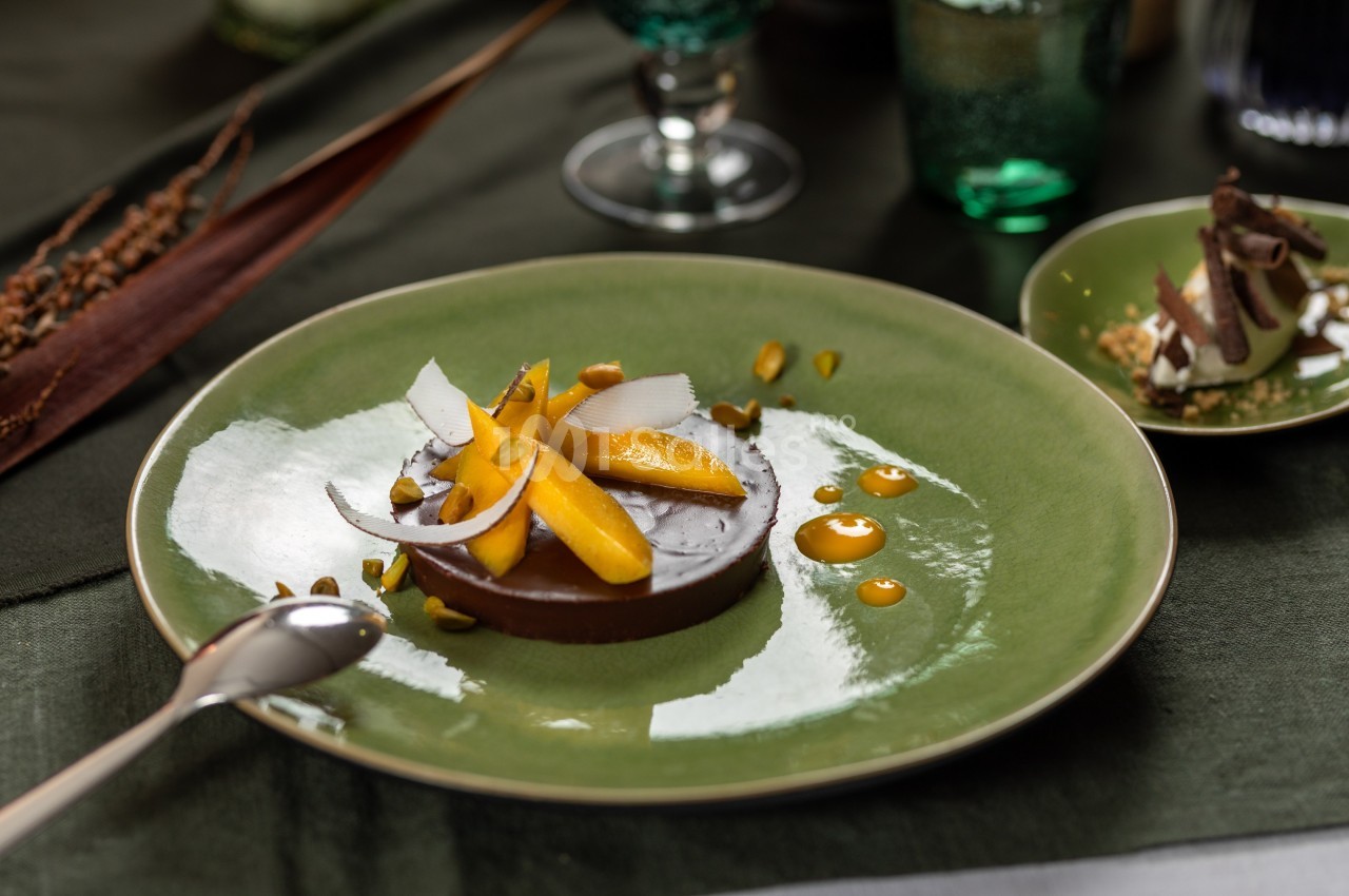 Dessert au chocolat garni de mangue, copeaux de noix de coco et pistaches, présenté sur une assiette verte.