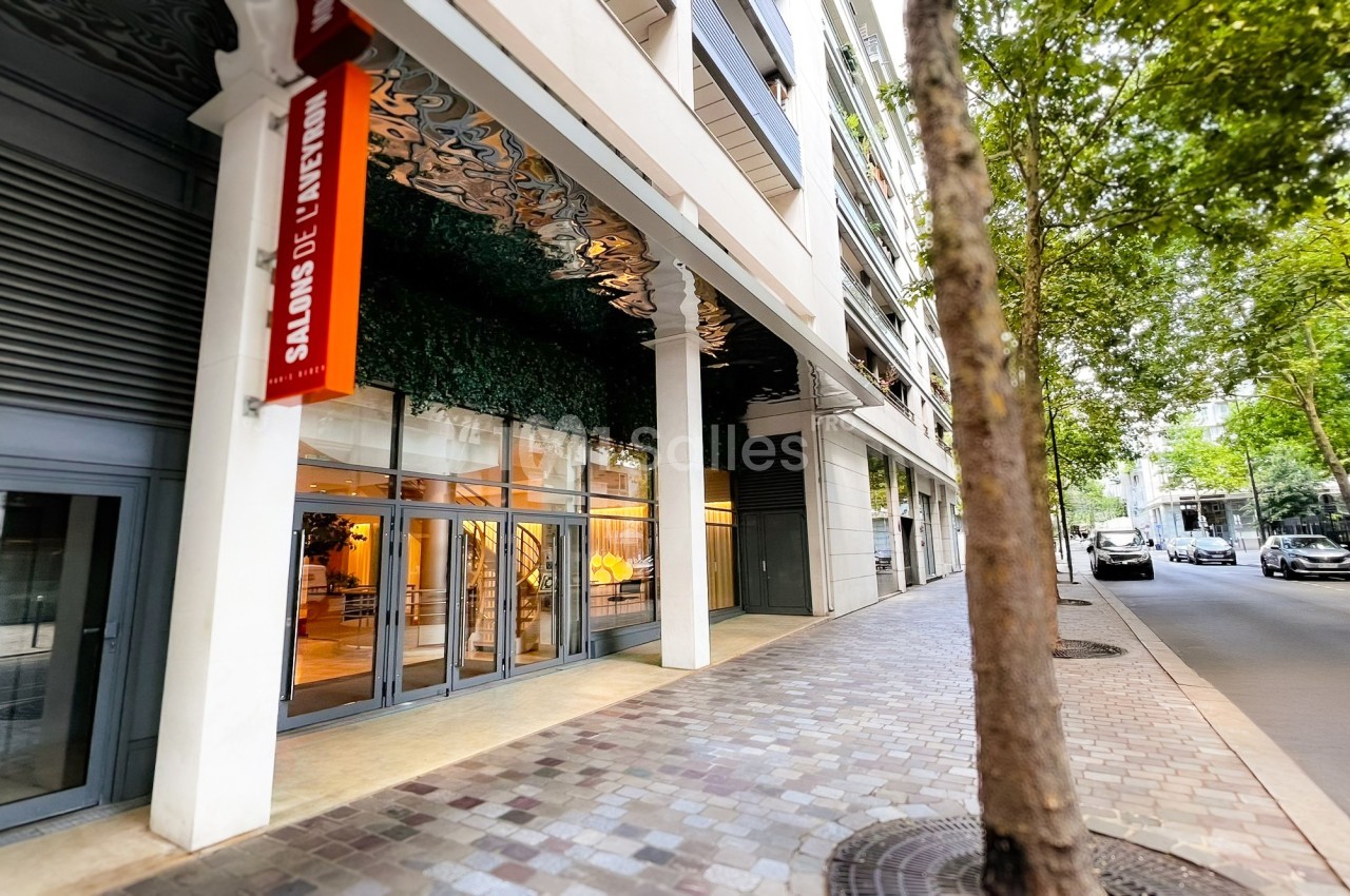 Façade moderne avec grandes baies vitrées et un panneau rouge indiquant ’Salons de l'Aveyron’, bordée d'arbres et d'une rue…