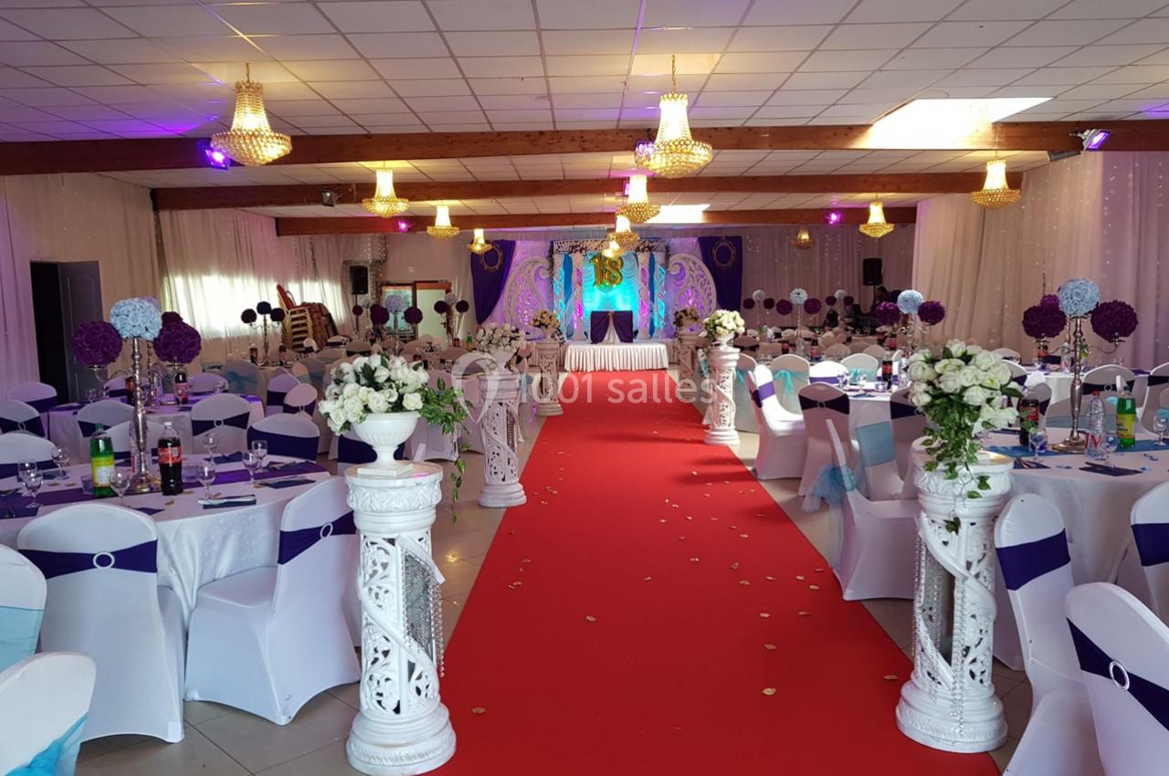 Salle de réception décorée avec des chaises blanches, des tables dressées, un tapis rouge et un éclairage coloré.