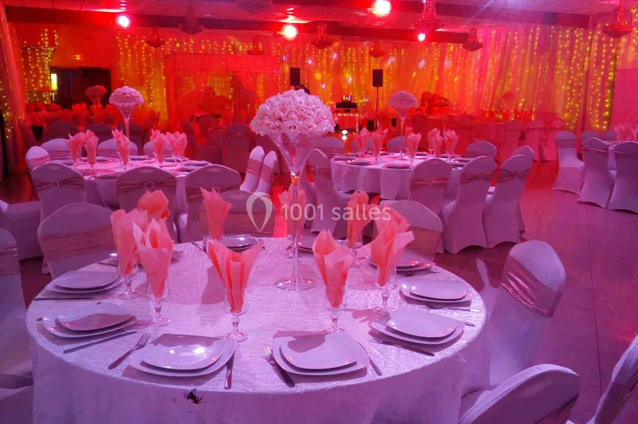 Salle de réception décorée avec des tables rondes dressées, nappes blanches, serviettes roses et éclairage rouge.
