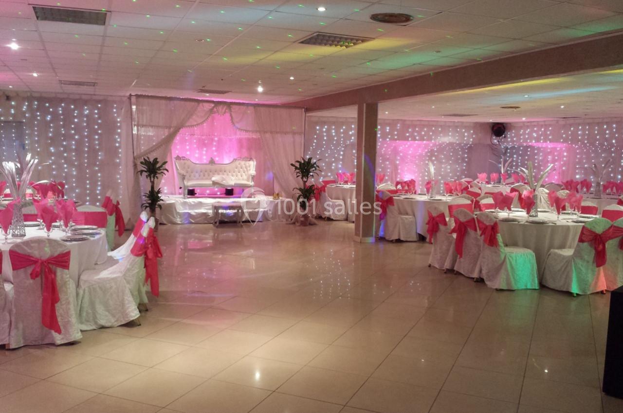 Salle de réception décorée avec des nappes blanches, des nœuds roses et un éclairage d'ambiance coloré.