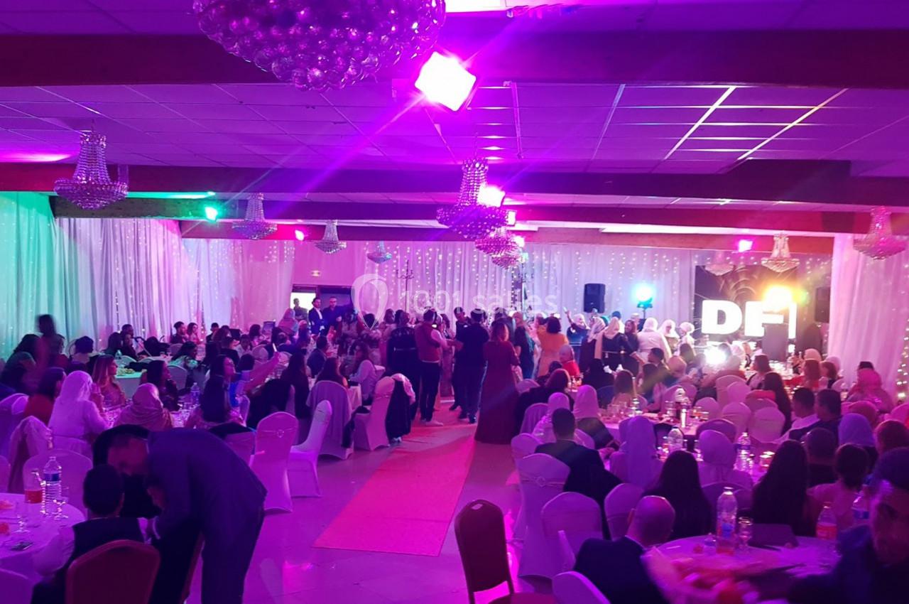 Salle de réception décorée avec des lumières colorées, remplie d'invités assis et debout lors d'un événement festif.