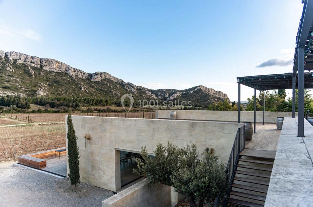 Bâtiment moderne en pierre entouré de vignes, avec une terrasse et des collines en arrière-plan sous un ciel dégagé.