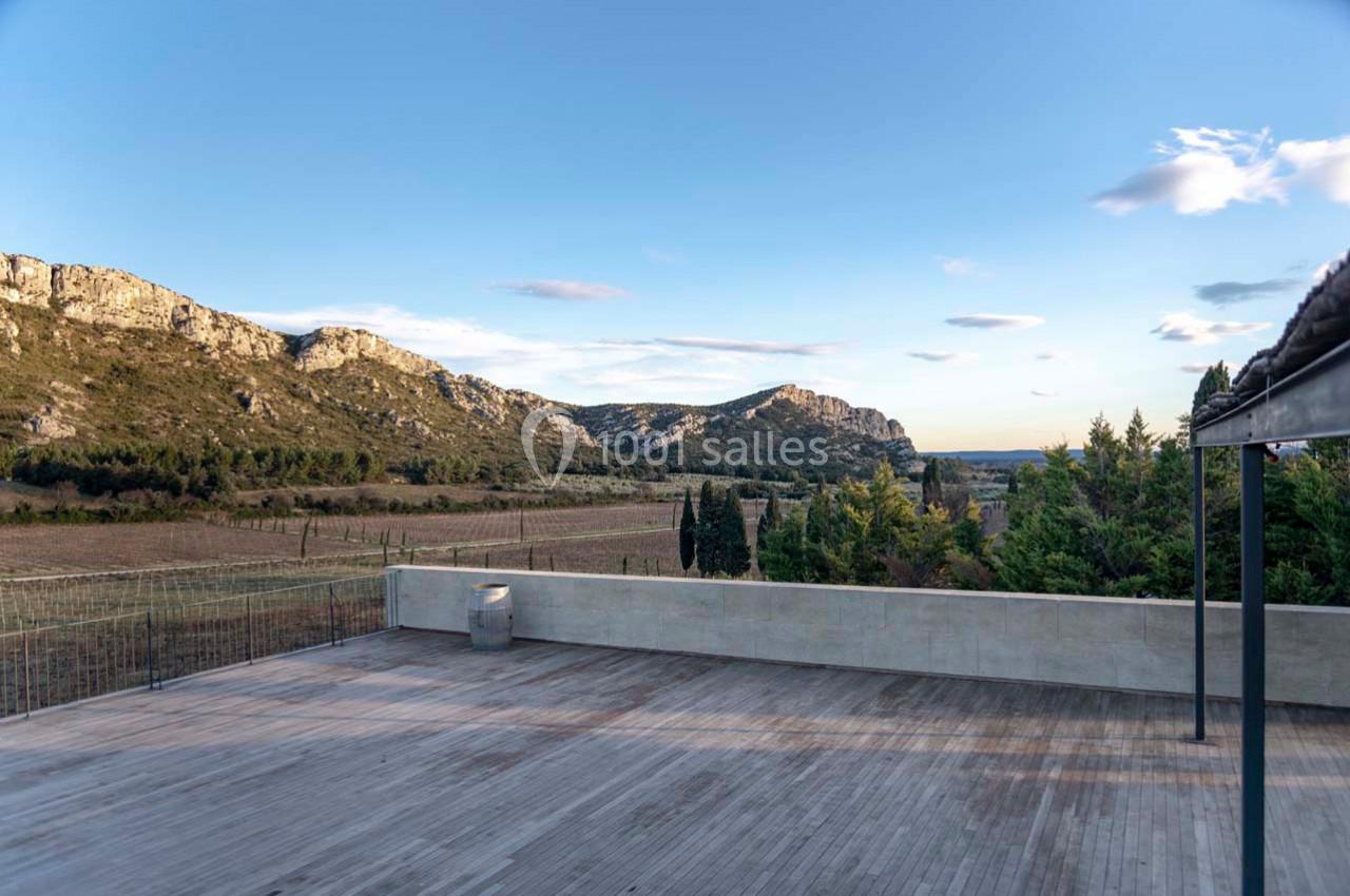Vue d'une terrasse en bois surplombant un paysage de collines, vignobles et végétation sous un ciel dégagé.