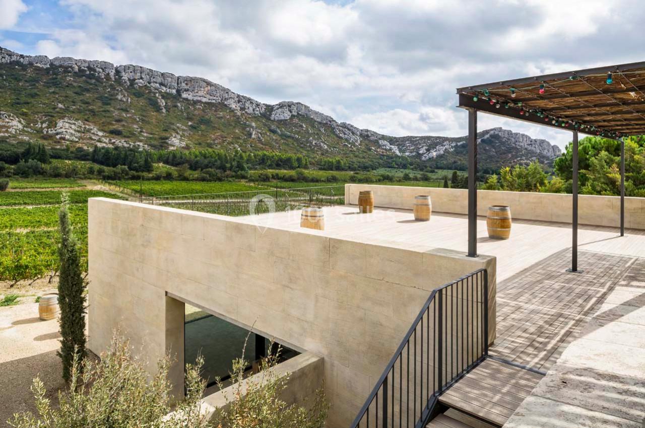 Terrasse en bois avec pergola surplombant un paysage de vignes et collines rocheuses sous un ciel partiellement nuageux.
