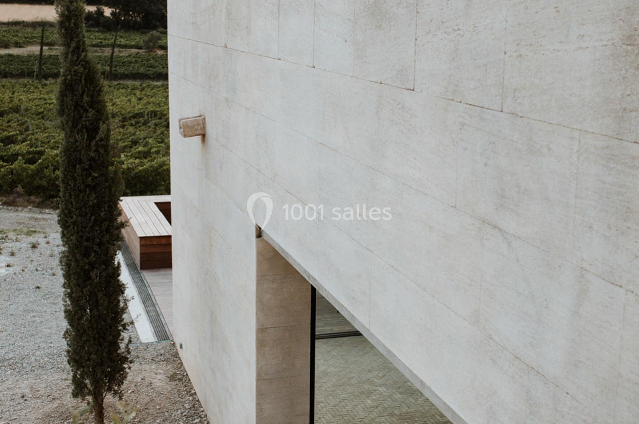 Façade moderne en pierre beige avec une grande fenêtre, un arbre élancé et un paysage rural en arrière-plan.