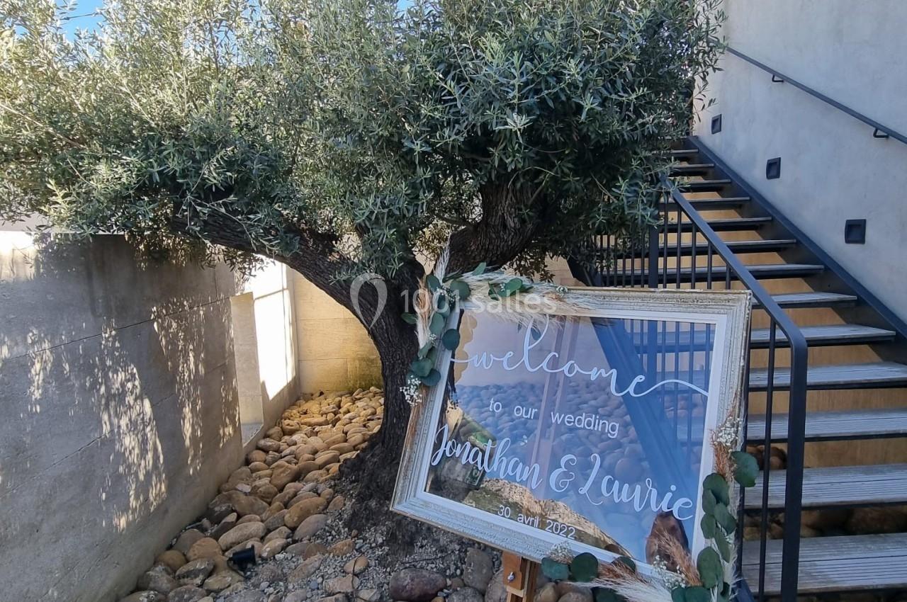 Panneau de bienvenue pour un mariage, posé sur un chevalet devant un olivier et un escalier extérieur en bois.
