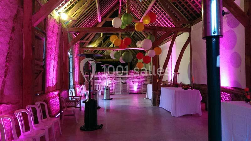 Salle de réception avec charpente en bois, éclairage coloré, lanternes suspendues et chaises blanches alignées.