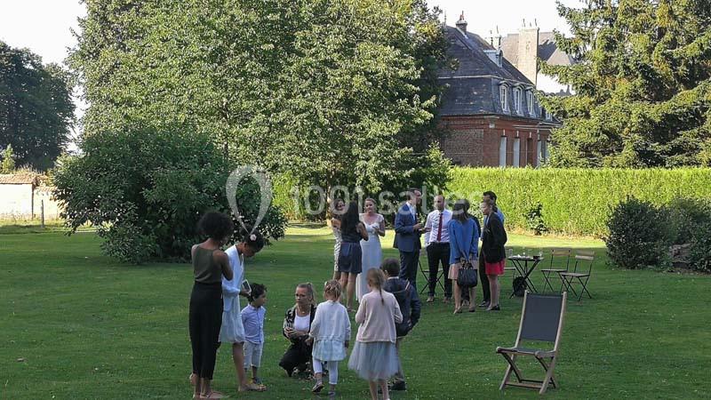 Un groupe de personnes, dont des enfants, discutent et jouent dans un jardin devant une maison en arrière-plan.