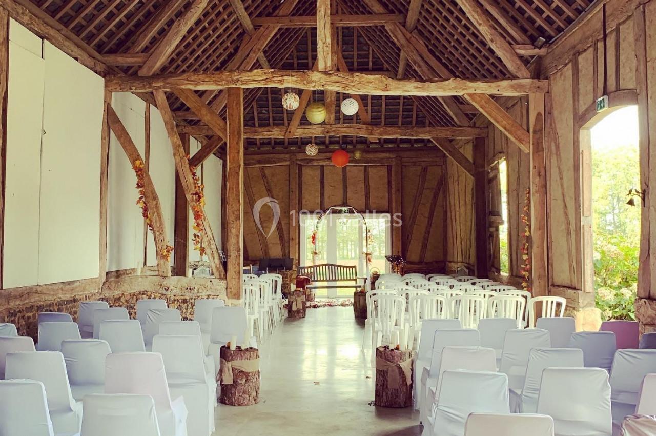 Salle de réception rustique avec poutres apparentes, chaises blanches alignées et décorations suspendues.