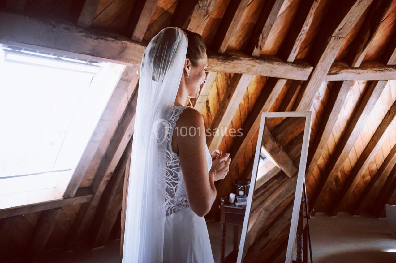 Une mariée en robe blanche et voile se prépare devant un miroir dans une pièce aux poutres apparentes.