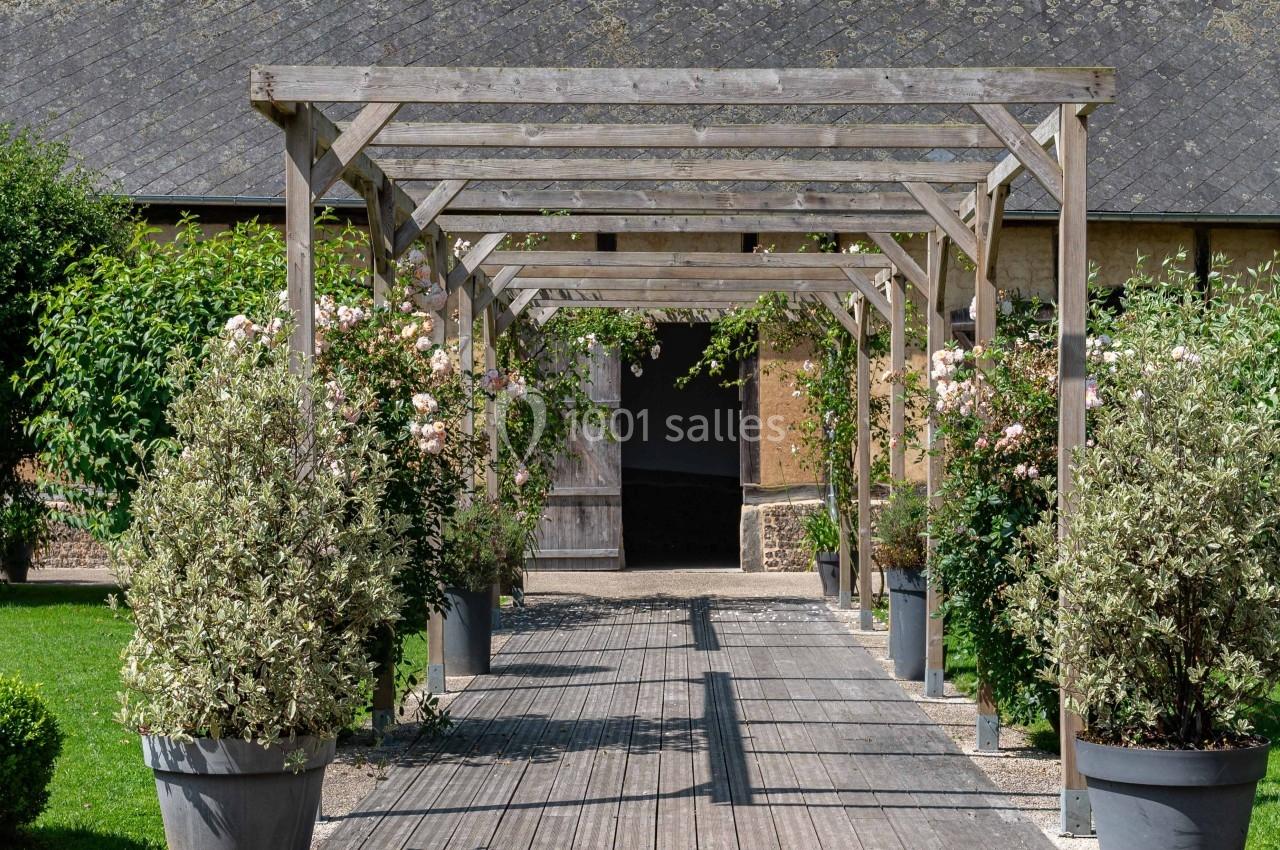 Allée en bois bordée de pots de plantes et de rosiers, menant à un bâtiment en pierre avec un toit en ardoise.