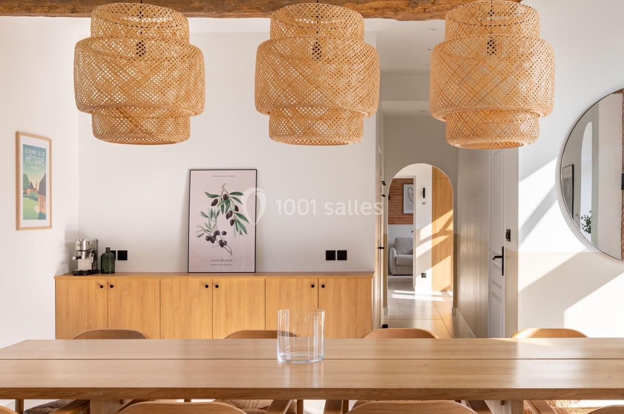 Salle à manger lumineuse avec table en bois, chaises assorties et suspensions en rotin sous une poutre apparente.