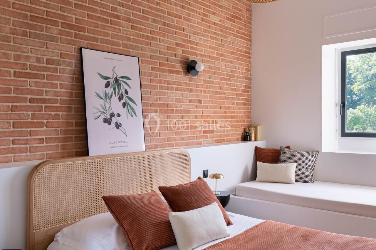 Chambre lumineuse avec mur en briques, lit aux tons terracotta, banquette près d'une fenêtre et suspension en rotin.