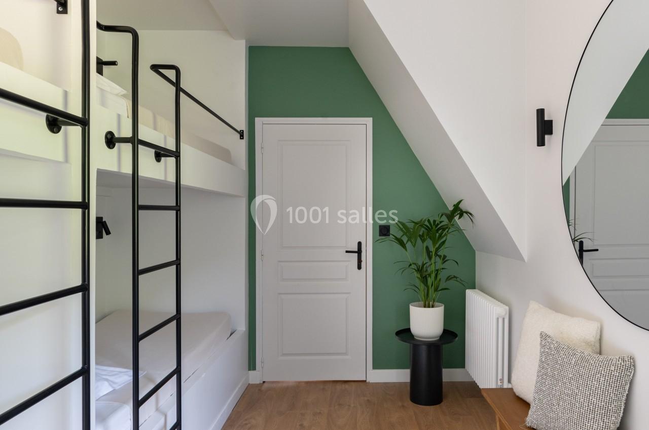 Chambre lumineuse avec lits superposés, mur vert, plante en pot, miroir rond et banc en bois sur un sol en parquet.