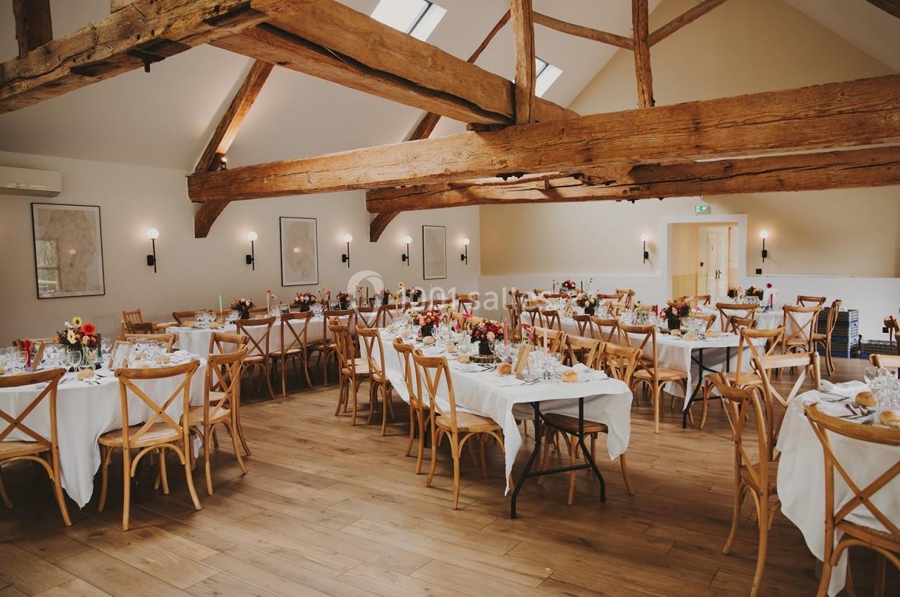 Salle de réception lumineuse avec poutres apparentes, tables décorées de fleurs et nappes blanches.