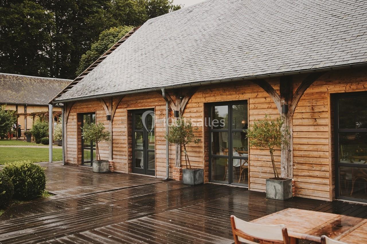 Façade d'un bâtiment en bois avec de grandes fenêtres, entouré de verdure et d'une terrasse mouillée par la pluie.