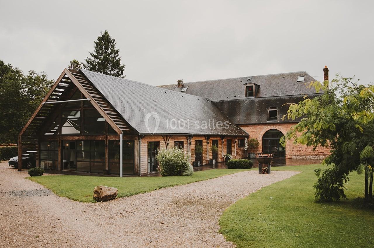 Bâtiment en briques et bois avec toit en ardoise, entouré de pelouse et d'arbres, sous un ciel nuageux.
