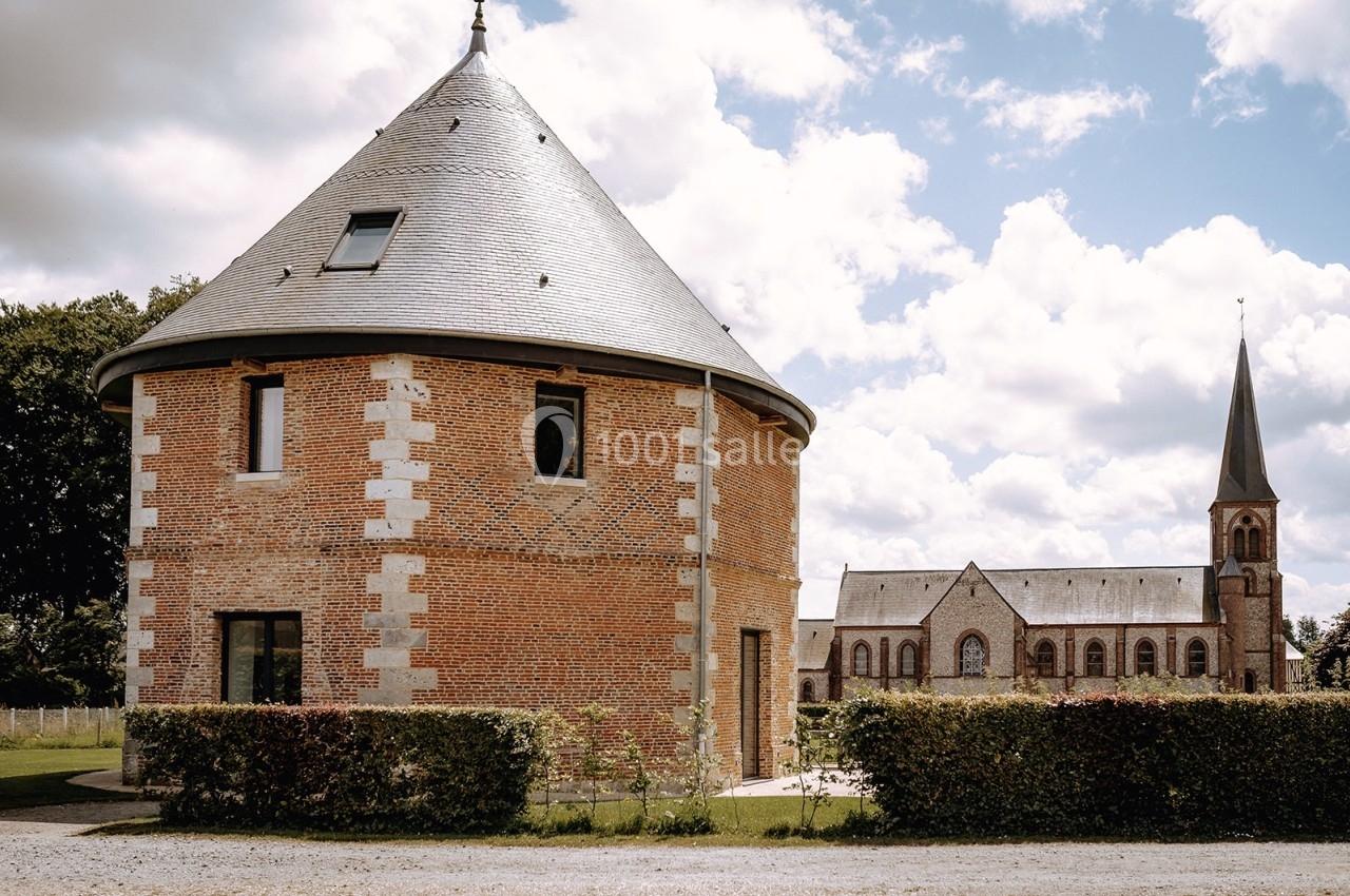 Bâtiment circulaire en briques avec toit en ardoise, situé près d'une église en pierre sous un ciel partiellement nuageux.